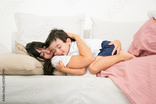 Brunette mother sleeping on couch and embracing cute son. Lovely little boy hugging young pretty mom, dreaming on sofa and smiling. Family, love and happiness concept