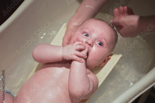 Infant baby boy washed indoors in small bath tub, mother holding and washing his head, view from above
