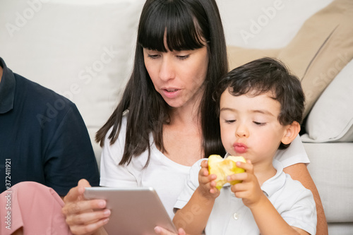 Adorable little boy eating fresh apples, sitting on mom lap while she using tablet. Communication and family leisure time concept