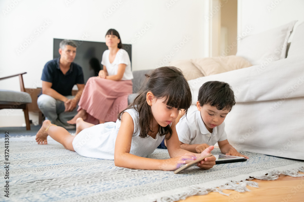 Cute little brother and sister using learning apps on gadgets, lying on floor while parents sitting together in background. Internet communication or digital technology concept