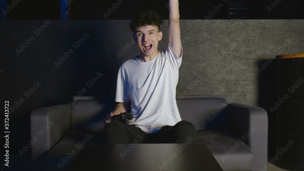 Funny excited young young man gamer winner holding joystick controller playing videogame sitting on sofa feeling overjoyed celebrating victory winning in video game alone at home