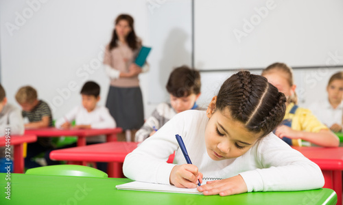 Portrait of little assiduous girl with pen and notebook at lesson in elementary school..