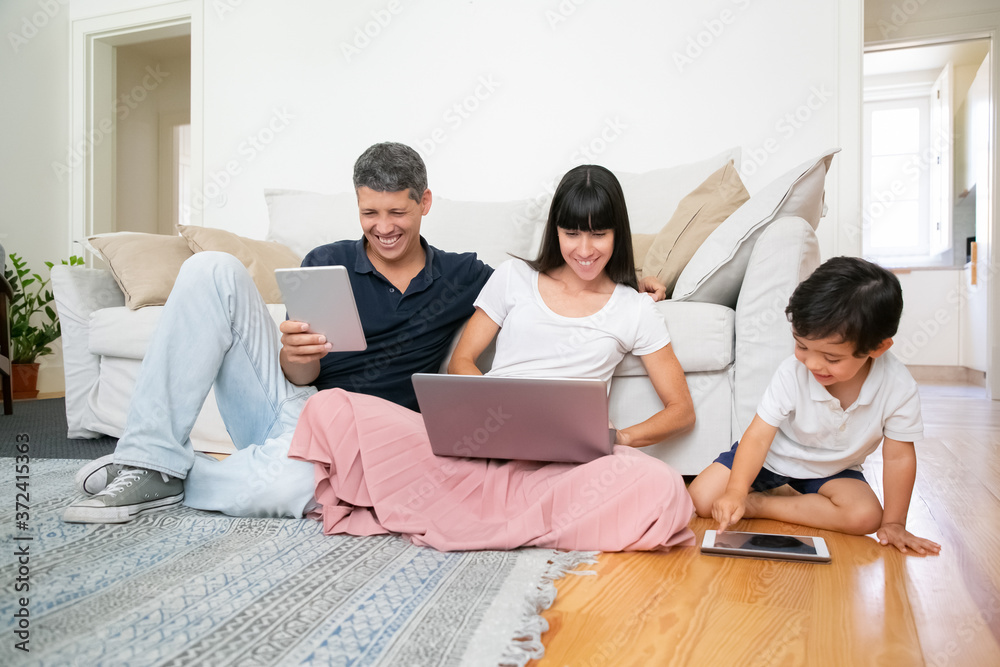 Cheerful family couple and cute boy sitting on apartment floor, laughing and using digital gadgets. Family leisure time and internet concept Cheerful family couple and cute boy sitting on apartment floor, laughing and using digital gadgets. Family leisure time and internet concept