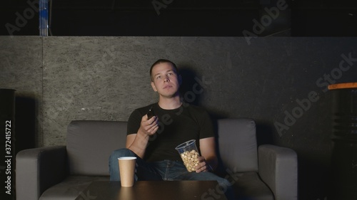 Handsome man eating popcorn and watching TV at home using remote control relaxing on couch
