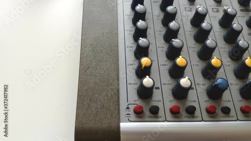 Shot of a side part of a mixing desk. A hand gets into the shot and adjusts the levels turning the different rotary knobs and presses a few buttons. photo