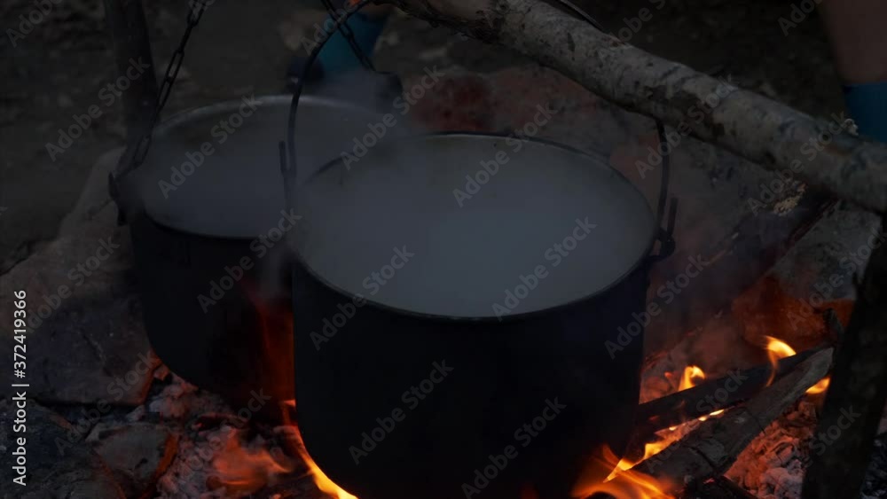 A two pots on a campfire at dusk. Male hand is pour boiling water from one pot to another A two pots on a campfire at dusk. Male hand is pour boiling water from one pot to another