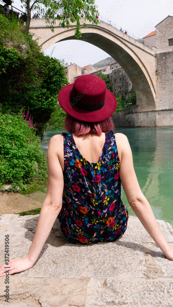 Caucasian woman in a floral dress with a hat sitting by the river Neretva and looks at the Old Bridge, Mostar, Bosnia and Herzegovina.