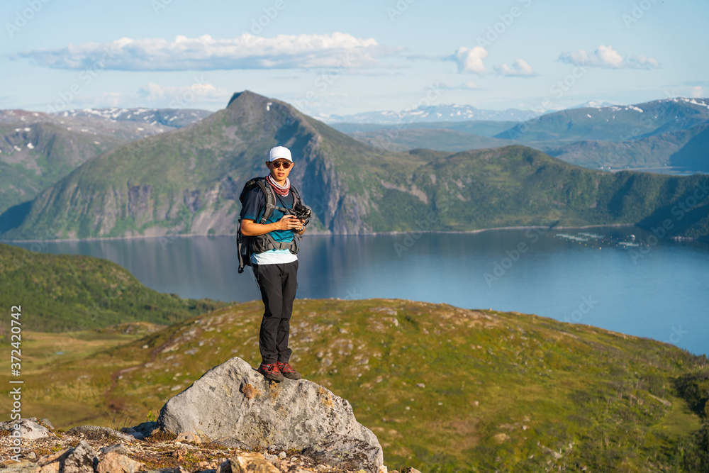 Young Asian man traveller with backpack and camera standing on rock at Senja island in summer season, Norway, Scandinavia