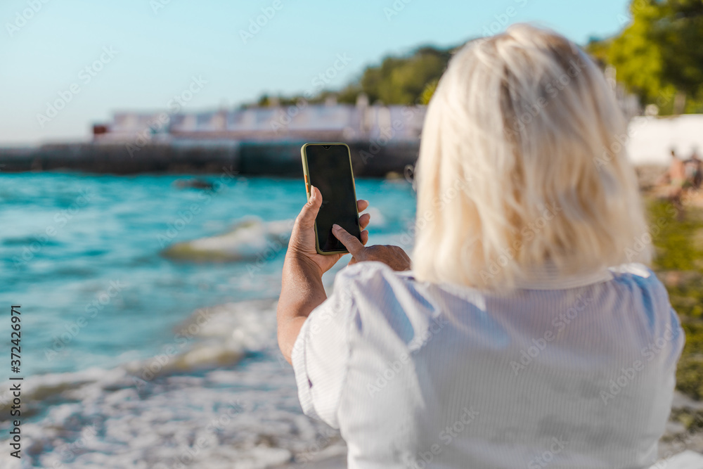 Blond woman back makes a photo on the beach of the sea by smartphone, enjoys leisure and summer vacation