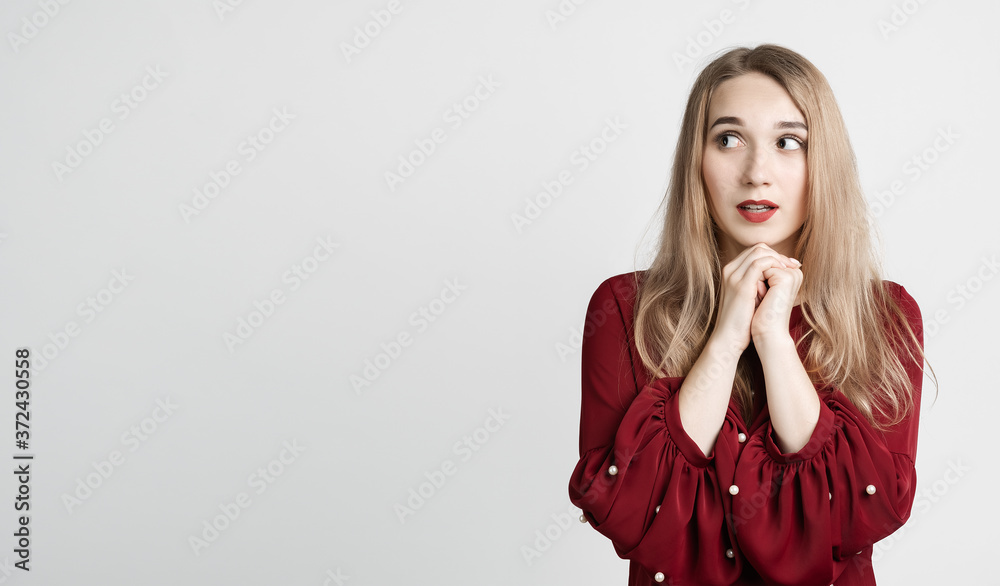 Pensive young woman with long blonde hair keeps hands together near chin, wears red blouse. Copy space Pensive young woman with long blonde hair keeps hands together near chin, wears red blouse. Copy space