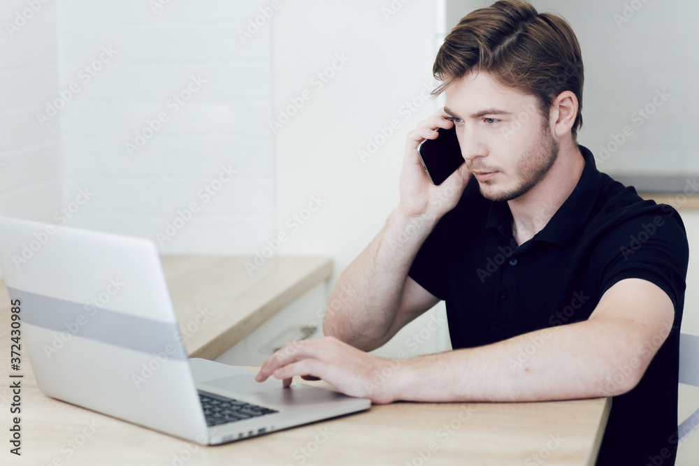 Young man working at home on a laptop talking on a cell phone, sitting at desk. Work at a distance during quarantine of coronavirus. Stay at home, self-isolation Young man working at home on a laptop talking on a cell phone, sitting at desk. Work at a distance during quarantine of coronavirus. Stay at home, self-isolation