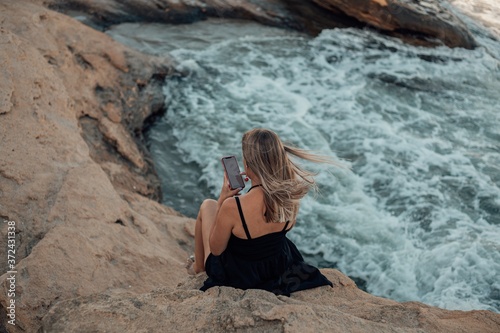 A young woman sits on the rocks near the sea and looks at the phone. View from above.