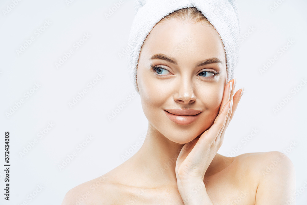 Beautiful female model with makeup, touches cheek, looks aside, has fresh clean skin after taking bath, wears soft towel on head, isolated over white background, copy space. Face care, hygiene concept Beautiful female model with makeup, touches cheek, looks aside, has fresh clean skin after taking bath, wears soft towel on head, isolated over white background, copy space. Face care, hygiene concept