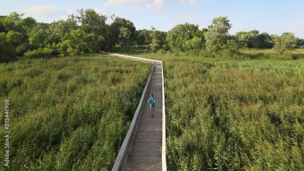 Drone following a young healthy man,running on the wooden path amidst lush greenery at old Cedar Avenue, Bloomington Minnesota. Aerial. Drone following a young healthy man,running on the wooden path amidst lush greenery at old Cedar Avenue, Bloomington Minnesota. Aerial.