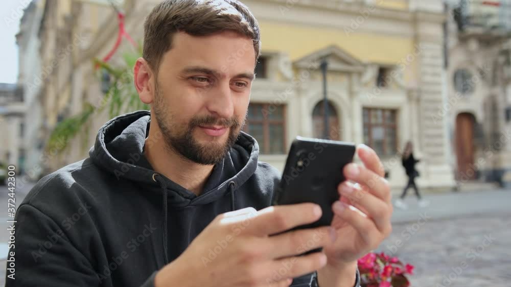 Handsome male student using smartphone. Smiling man making video call via mobile phone in hand. Concept of guy internet-enabled electronic device outdoor. Handsome male student using smartphone. Smiling man making video call via mobile phone in hand. Concept of guy internet-enabled electronic device outdoor.