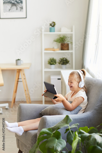 Girl with down syndrome sitting on armchair in headphones watching something on tablet pc and laughing
