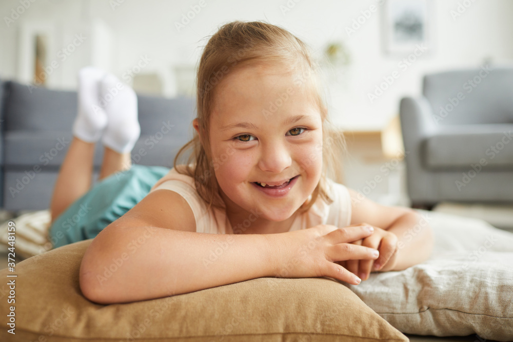 Portrait of little girl with down syndrome smiling at camera while lying on pillows on the floor