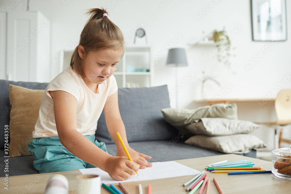 Girl with down syndrome sitting at the table and drawing picture with colorful pencils in the living room at home Girl with down syndrome sitting at the table and drawing picture with colorful pencils in the living room at home