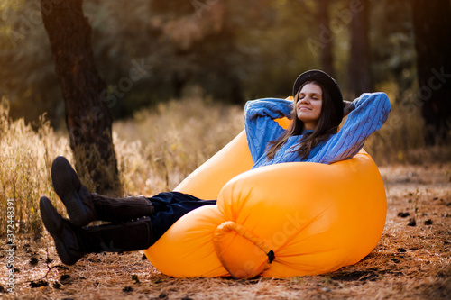 Happy autumn portrait of young woman in blue sweater and black hat lying on orange inflatable sofa outdoor in forest photo