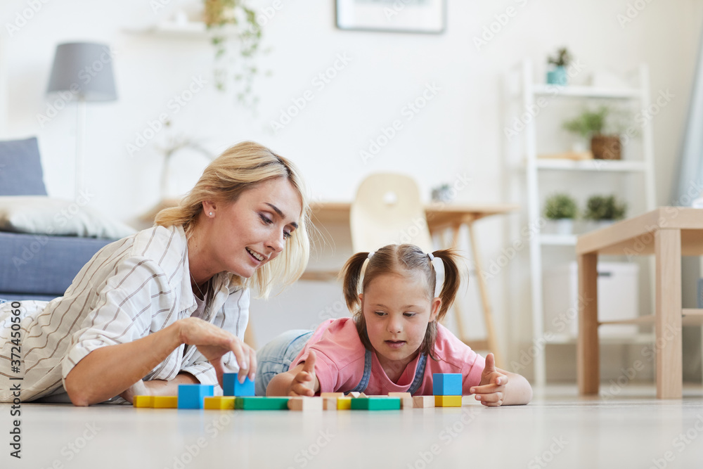 Young mother playing with her daughter with down syndrome in colored blocks while they lying on the floor in the room Young mother playing with her daughter with down syndrome in colored blocks while they lying on the floor in the room