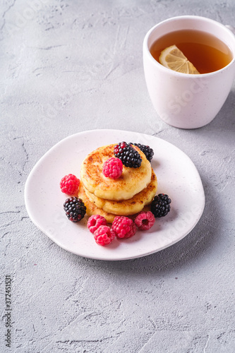Cottage cheese pancakes, curd fritters dessert with raspberry and blackberry berries in plate near to hot tea cup with lemon slice on stone concrete background, angle view