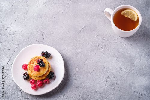 Cottage cheese pancakes, curd fritters dessert with raspberry and blackberry berries in plate near to hot tea cup with lemon slice on stone concrete background, angle view copy space