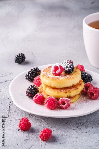 Cottage cheese pancakes and powdered sugar, curd fritters dessert with raspberry and blackberry berries in plate near to hot tea cup with lemon slice on stone concrete background, angle view