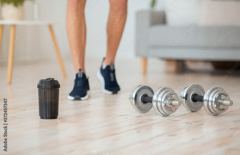 Guy in sneakers goes to dumbbells and glass of protein shake on wooden floor