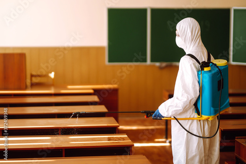 Worker wearing protective suit sprays disinfectant school class. COVID-19.