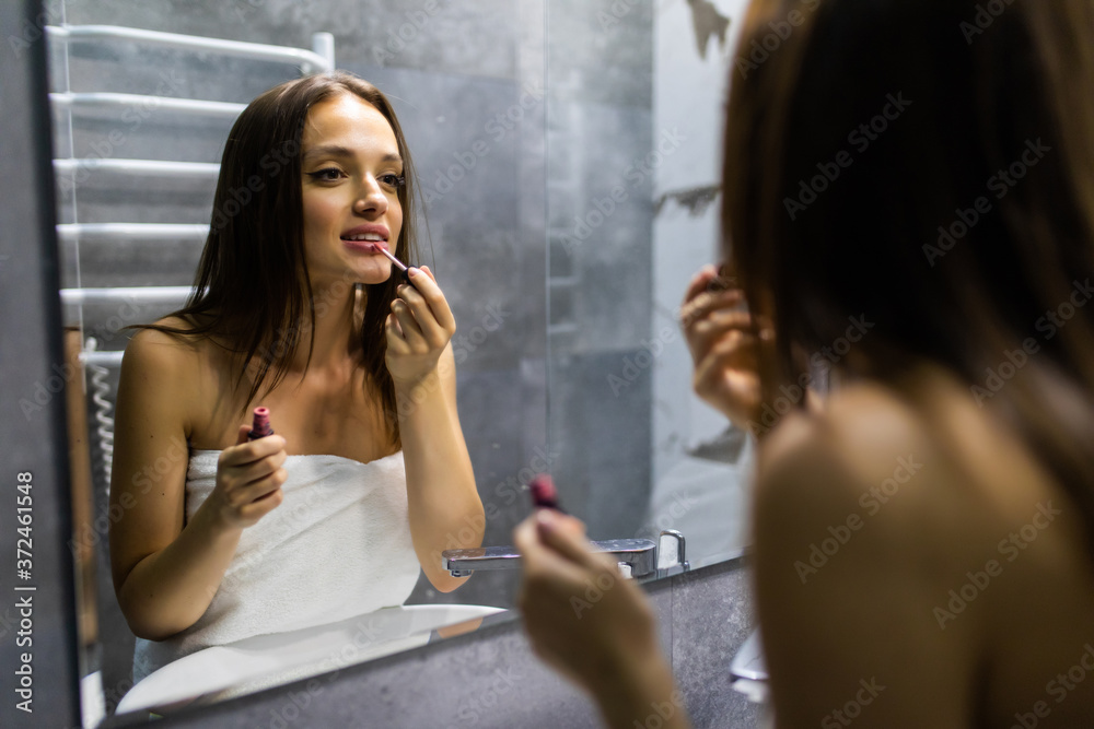 Beautiful millennial girl in towel look in mirror get reading in morning apply red lipstick. Caucasian young woman do makeup paint lips after shower in home bathroom. Beauty care routine concept Beautiful millennial girl in towel look in mirror get reading in morning apply red lipstick. Caucasian young woman do makeup paint lips after shower in home bathroom. Beauty care routine concept