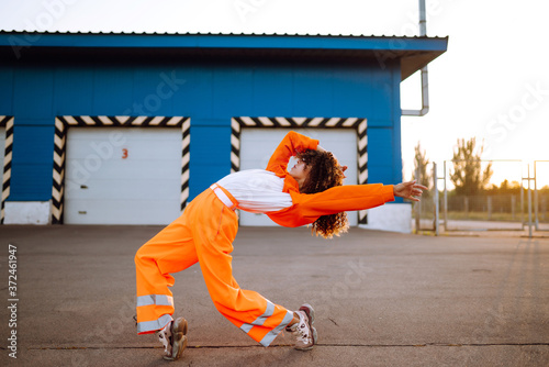 Young African American woman - dancer dancing in the street at sunset. Stylish woman with curly hair in an orange suit  showing some moves. Sport, dancing and urban culture concept.