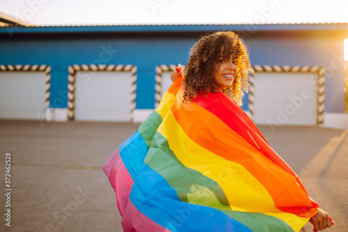 Young African American lesbian woman with LGBT rainbow flag in the street at sunset. Stylish woman with curly hair in an orange suit posing with lgbt pride flag.