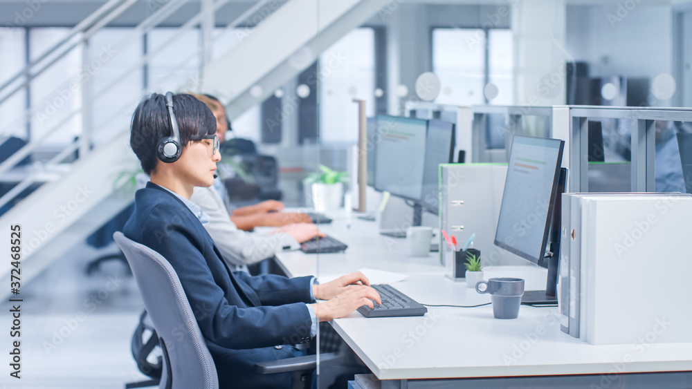 Shot of Handsome and Beautiful Asian Customer Service Operator Working at His Desk Personal Computer in a Busy Modern Call Center. He is Wearing Headsets and Actively Taking Calls. Shot of Handsome and Beautiful Asian Customer Service Operator Working at His Desk Personal Computer in a Busy Modern Call Center. He is Wearing Headsets and Actively Taking Calls.