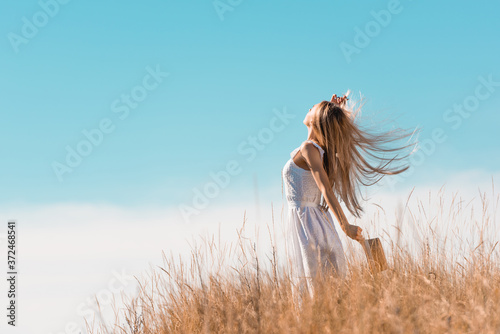 selective focus of blonde woman in white dress holding straw hat while standing with raised hand on grassy hill