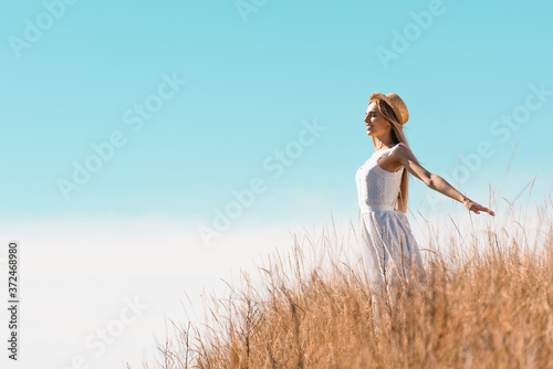 blonde woman in straw hat and white dress standing with outstretched hands on hill against blue sky