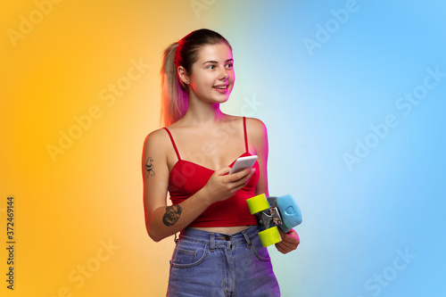 Skatergirl, scrolling phone. Caucasian young woman's portrait on gradient studio background in neon. Beautiful female model in casual style. Concept of human emotions, facial expression, youth, sales © master1305