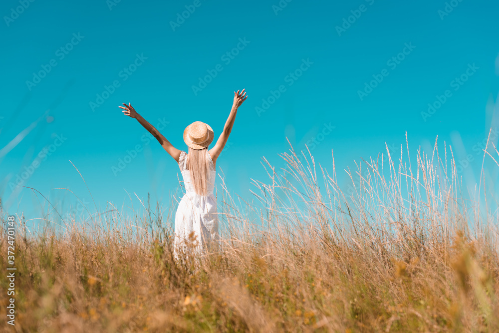 back view of woman in straw hat and white dress standing with raised hands on grassy meadow, selective focus back view of woman in straw hat and white dress standing with raised hands on grassy meadow, selective focus