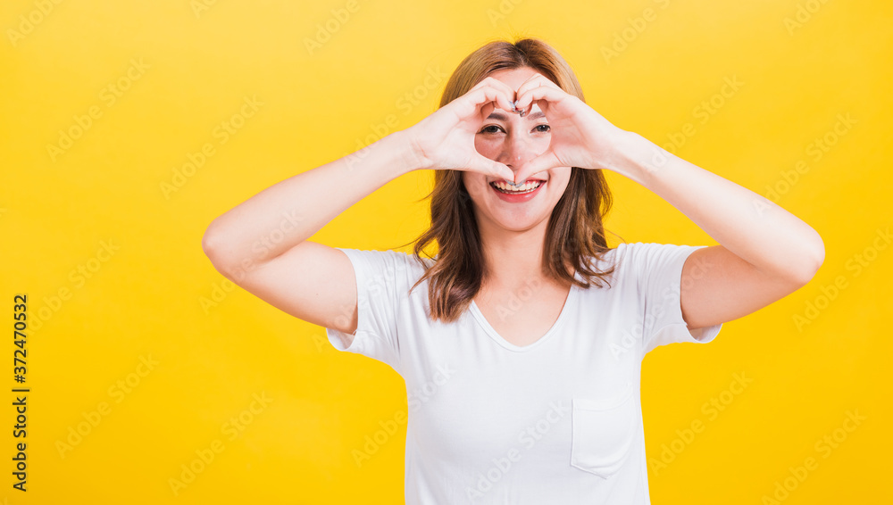 Fototapeta premium Azjatycki tajski szczęśliwy portret piękna śliczna młoda kobieta uśmiech stojąca zrobić palec serce figura symbol kształt znak z dwiema rękami i patrząc kamery, studio strzał izolowane żółte tło z miejsca na kopię