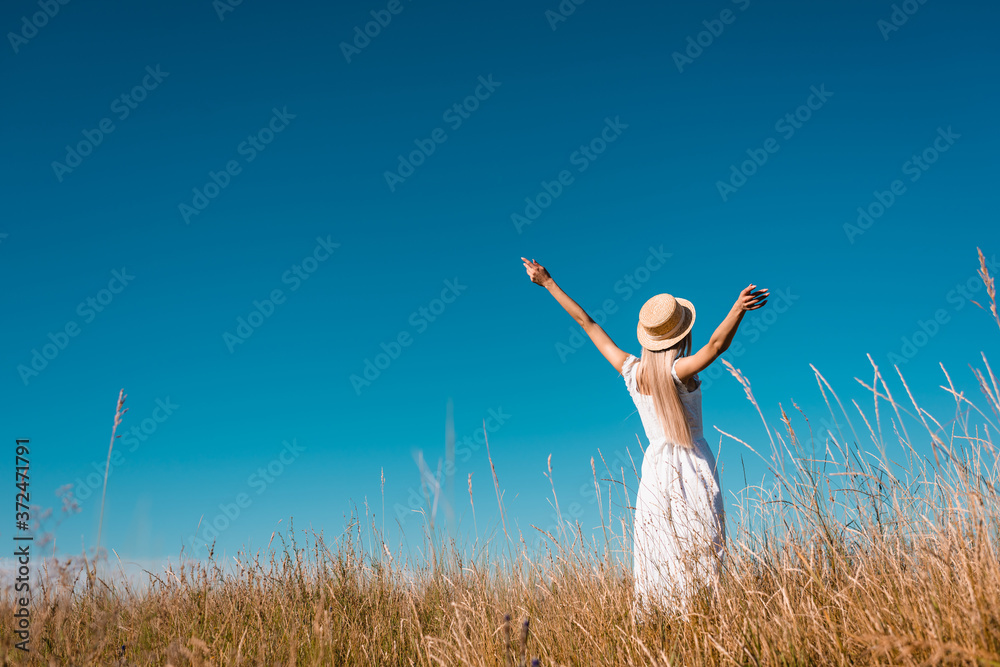 back view of woman in white dress and straw hat standing with outstretched hands in grassy field against blue sky back view of woman in white dress and straw hat standing with outstretched hands in grassy field against blue sky