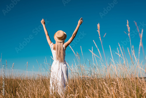 back view of stylish woman in white dress and straw hat standing in grassy meadow with raised hands against blue sky