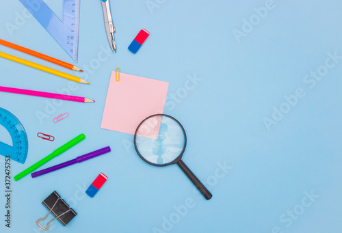 The concept of school and students. An empty square colored paper for notes in the middle, a magnifying glass next to it. School and office stationery on the edges on a blue background. Copyspace