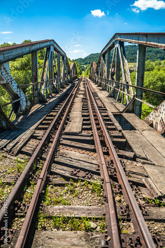 Railway bridge over river in Polish mountain. Heavy rusty steel old industrial train overpass in natural scenic landscape, blue summer sky.