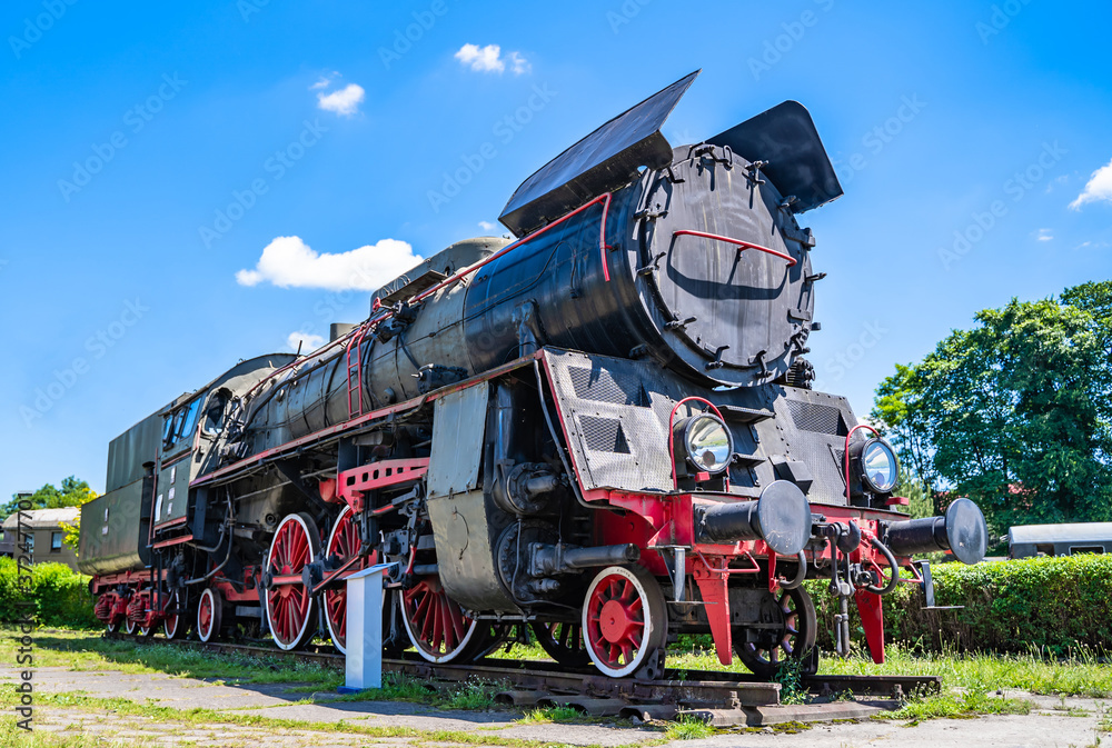Naklejka premium Huge vintage steam locomotive, red painted steel wheel detail close up. Coal-powered steam train stands on a siding. Classic gigantic heavy railway machinery. Side view of power parts of machine.