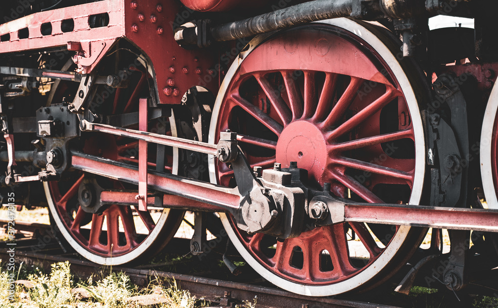 Huge vintage steam locomotive, red painted steel wheel detail close up. Coal-powered steam train stands on a siding. Classic gigantic heavy railway machinery. Side view of power parts of machine.