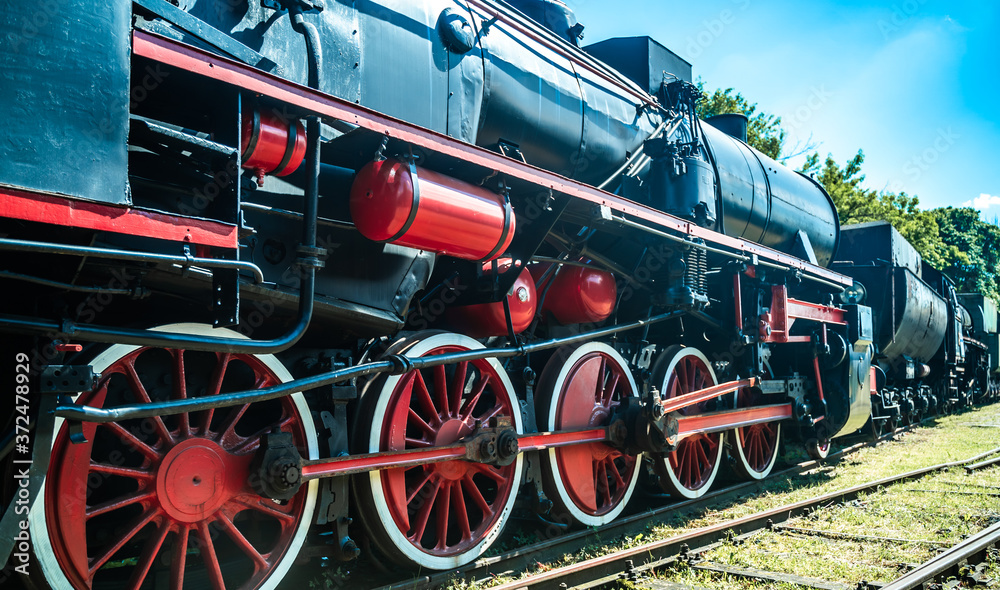 Naklejka premium Huge vintage steam locomotive, red painted steel wheel detail close up. Coal-powered steam train stands on a siding. Classic gigantic heavy railway machinery. Side view of power parts of machine.