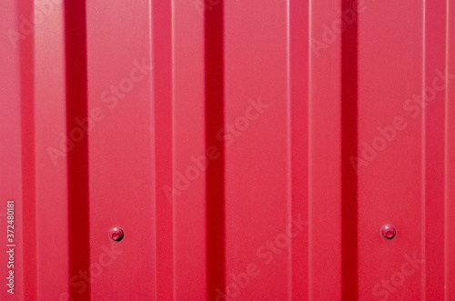A fragment of a red metal profile of the Corrugated galvanised iron type attached to the base by roof screws with a pressure washer and a hexagon head.  Used for covering roofs and fences.
