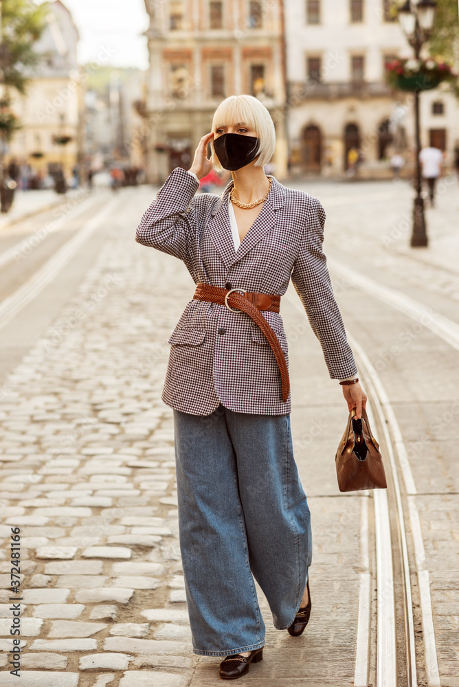 Street style during quarantine: woman wearing protective face mask, trendy autumn blazer, wide leg jeans, walking in street of European city