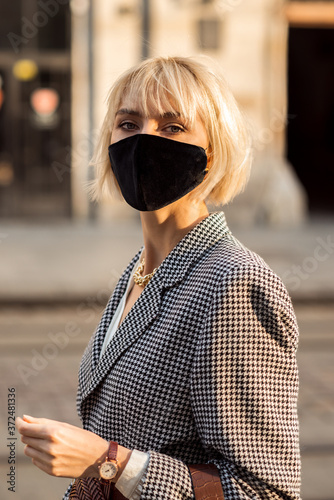 Street style during quarantine: elegant blonde woman wearing black protective face mask, trendy autumn blazer, brown wrist watch, posing in street of European city