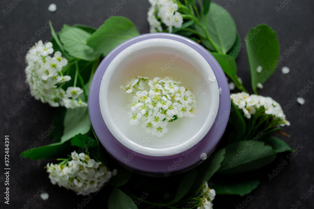 Close up of lilac jar with white cream of delicate texture, inside which-white flowers. Spirea twigs with flowers and green leaves are lying around. Dark background