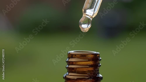 Macro shot essential oil dripping from a pipette on blurry natural background. Extreme close up hand taking transparent oil from a brown glass bottle